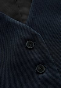 Marineblauw corduroy stof met een gestructureerd ontwerp, twee zwarte knopen en een contrasterende zwarte voering. Close-up van het materiaal en de details.