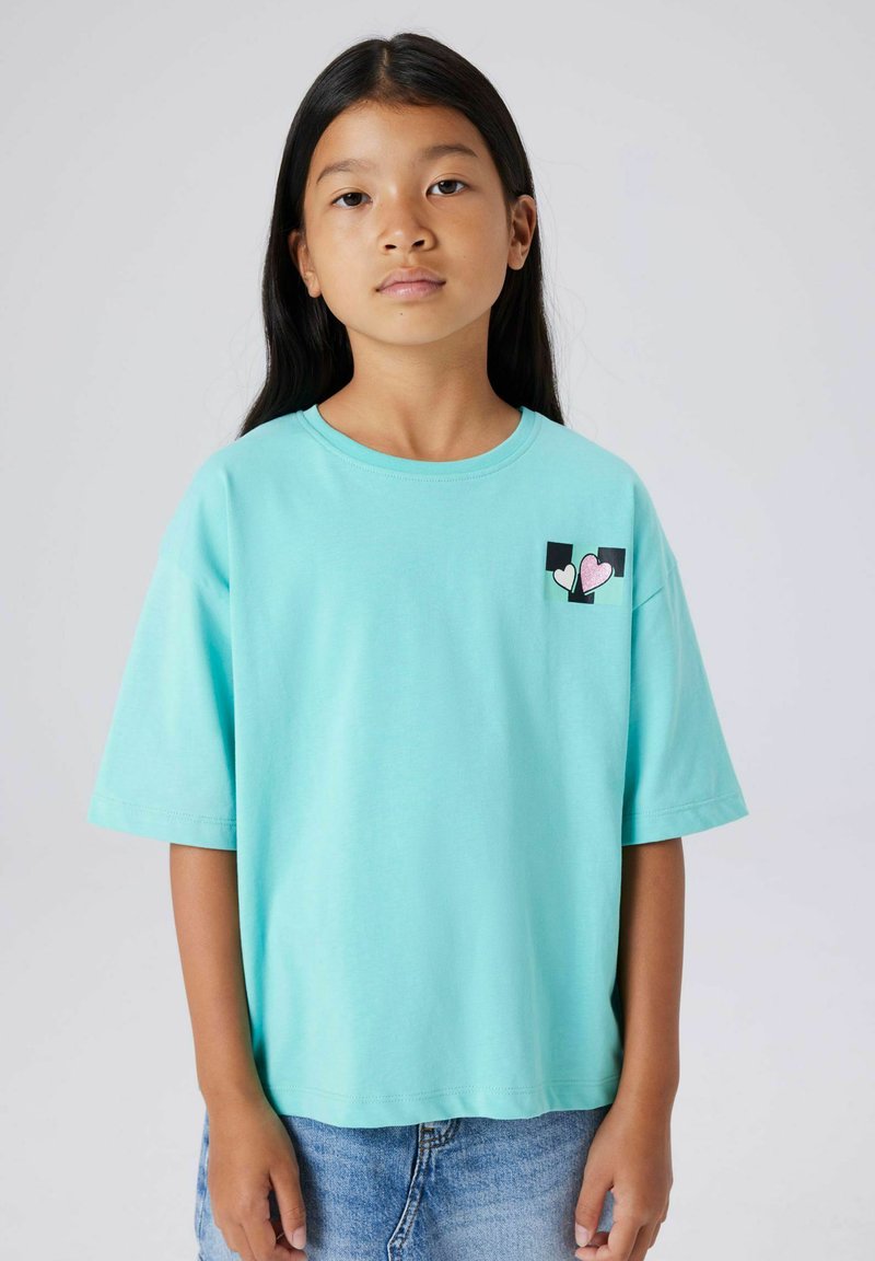 Camiseta de algodón turquesa con mangas cortas, de corte holgado, con un gráfico de bolsillo con corazones en rosa y blanco, combinada con pantalones cortos de mezclilla.