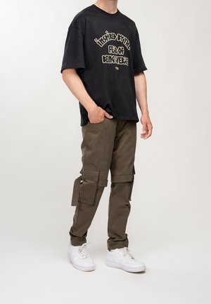 Homme portant un t-shirt noir à motif, un pantalon cargo marron avec poches latérales, et des baskets blanches, debout avec une main dans la poche.