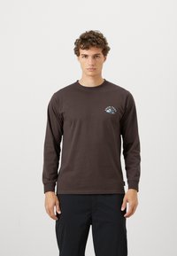 Long-sleeve, dark brown cotton T-shirt with a round neck and a small blue mountain graphic on the left chest. Smooth texture.