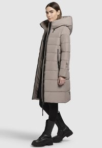 Párnázott hosszú kabát világos bézs színben, magas gallérral, teljes cipzárral és oldalsó zsebekkel. Fekete katonai csizmával és leggingsszel párosítva.