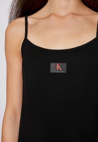 Černý top bez rukávů vyrobený z hladké tkaniny, s obdélníkovým logem s červeným "cK" a textem "Calvin Klein Jeans".