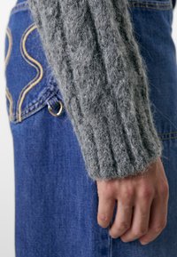 Manche de pull en tricot côtelé gris à côté d'une jupe en denim bleu avec des coutures dorées et un détail en métal en anneau sur la poche.