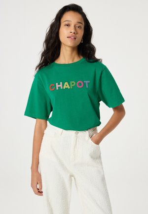 Woman with long dark hair wears green t-shirt with colorful "CHAPOT" text and white textured high-waisted pants, hand in pocket.