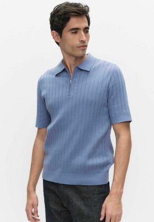 Mand iført en lyseblå kortærmet ribstrikket polotrøje med lynlås foran, kombineret med mørke jeans, kigger til siden.