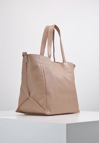 Beige läder tote-väska med dubbla axelband, något strukturerad rektangulär form och synliga sömmar, utställd på en vit yta.