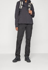 Czarna wodoodporna kurtka z kapturem, zestawiona z pasującymi czarnymi spodniami cargo z kieszeniami na zamek. Włożone z czarnymi butami trekkingowymi.