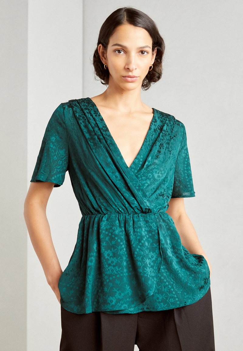 Blusa avvolgente turchese con maniche corte, tessuto strutturato con motivi floreali, scollatura a V profonda e vita arricciata con tasche laterali.
