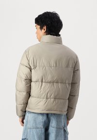 Puffer jacket in light gray with a high collar, quilted texture, and elastic cuffs. Worn over blue oversized denim pants.
