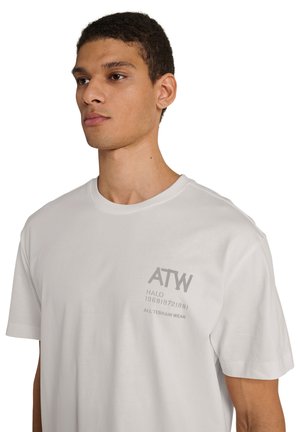 Ung mand iført en lysegrå T-shirt med teksten "ATW" og "ALL TERRAIN WEAR" på venstre bryst, vendt let mod venstre på en hvid baggrund.