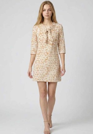 Joven mujer con vestido beige de patrón geométrico, con mangas tres cuartos y detalle de lazo en el escote, caminando hacia la cámara sobre un fondo liso.