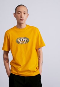 Giovane uomo con tatuaggi che indossa una maglietta gialla con un logo Adidas distorto, in piedi con le mani in tasca su uno sfondo neutro.