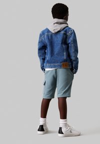 Jeansjacke mit einer grauen Kapuze, hellblaue Shorts mit Taschen und weißen Sneakers mit schwarzem Akzent. Aus Baumwolle, lockere Passform.
