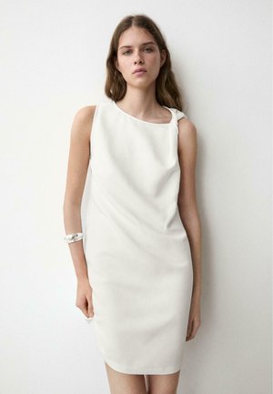 Junge Frau mit langen braunen Haaren, die ein ärmelloses weißes Kleid mit einem Knoten an einer Schulter und ein silbernes Armband am Arm trägt.