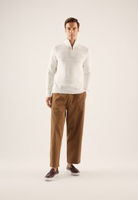 Suéter blanco de cremallera con patrón de tejido texturizado en los hombros, combinado con pantalones sueltos marrones y zapatos oscuros tipo slip-on.