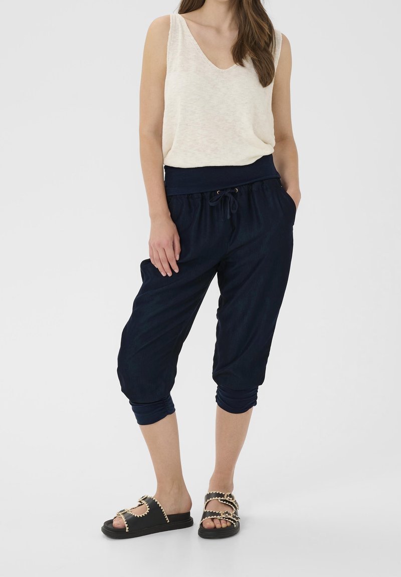 Femme portant un haut blanc sans manches, un pantalon court bleu marine avec cordon, et des sandales noires avec des détails en chaîne dorée, debout les mains dans les poches.