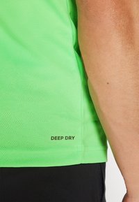 Neon grønn idretts skjorte laget av teksturert stoff med en "DEEP DRY"-etikett ved kanten, lagdelt over svarte shorts.