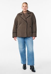 Brown, textured, oversized fleece jacket with a notched collar, black stitching details, and two front pockets, worn over denim jeans.