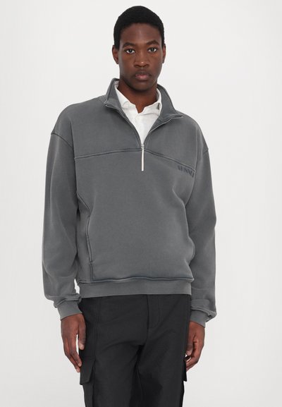 Sudadera de felpa gris con cuello de media cremallera, bolsillo y puños acanalados. Presenta detalles de costura sutiles y un pequeño logo en el pecho.