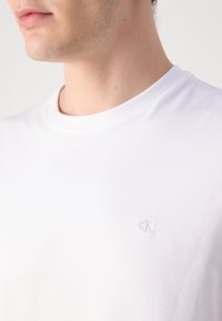 Gros plan d'une personne portant un t-shirt blanc uni à col rond avec un petit logo "cK" subtil brodé sur la poitrine.