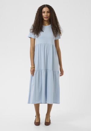 Naine, kellel on pikad lokkis juuksed, seljas hele sinine lühikeste varrukatega midi kleit ja pruunid punutud kontsad, seisab ühevärvilise tausta ees.