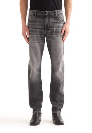 Mann trägt verblasste schwarze gerade geschnittene Jeans, schwarzes Hemd und schwarze Stiefel, steht mit entspannten Händen an den Seiten.