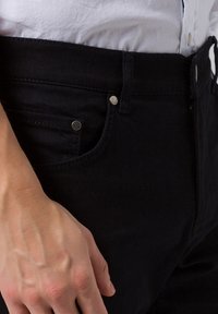 Černé džíny z denimu mají slim fit, kovové knoflíkové detaily a hladkou texturu. Detailní pohled na pas a ruku držící látku.