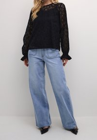 Haut à manches longues en dentelle noire avec un col rond et des poignets évasés, associé à un jean large bleu clair et des chaussures noires à bout pointu.