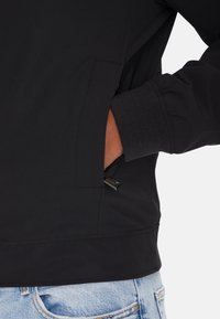 Giacca nera con una texture liscia, dotata di tasche laterali, polsini elastici e apertura con zip all'altezza dell'anca. Design casual con linee pulite.