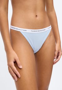 Frau trägt einen hellblauen Bikini-Unterwäsche-Stil mit weißem Bund, auf dem das Tommy Hilfiger Logo zu sehen ist, vor einem einfarbigen hellen Hintergrund.