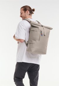 Mochila de roll-top en gris claro, fabricada en material impermeable. Cuenta con correas ajustables, un bolsillo frontal con cremallera y tiradores de cremallera coloridos.