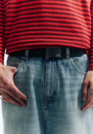 Persoană purtând o cămașă cu dungi roșii și negre băgată într-o pereche de blugi albaștri decolorați cu o curea neagră, mâinile sprijinite lângă buzunare.