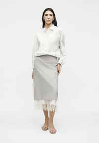 Γυναίκα που φοράει ένα λευκό ριγέ πουκάμισο και γκρι φούστα μέσης με δαντέλα στο τελείωμα, στέκεται μπροστά σε ένα απλό φωτεινό φόντο.