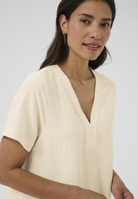 Crème claire, haut texturé avec un col en V et des manches courtes, présentant une finition en dentelle décorative le long de l'encolure. Design simple et décontracté.