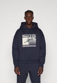 Μπλε ναυτικού βαμβακερή φούτερ με γραφικό τύπωμα που απεικονίζει το "Monte Carlo" και τη σιλουέτα ενός τενίστα. Χαλαρή εφαρμογή με τσέπη καγκουρό.