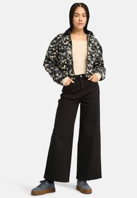 Zwart doorbloemd pufferjack, beige top, zwarte wijde broek en grijze schoenen met bruine zolen. Robuuste texturen en opvallende kleuren.