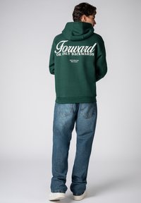Dunkelgrüner Hoodie mit einem großen weißen Aufdruck mit dem Text „Forward OR ONLY BACKWARDS“ auf der Rückseite, kombiniert mit hellblauen Jeans.