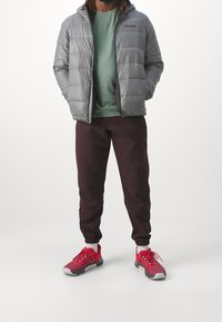 Grau gefütterte Jacke mit Kapuze, grünes Shirt, bordeauxrote Jogginghose und rote Sneakers mit grauen Akzenten. Texturierte Materialien und lässige Passform.