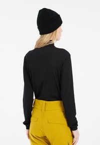 Zwarte blouse met lange mouwen en hoge hals, gecombineerd met gele hoge taillebroek met een gladde textuur en pocketdetails. Zwarte beanie op het hoofd.