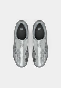 Ezüst színű bebújós cipők sima, fényes fémes felülettel és finom hullámmintákkal. Fekete talpbéléssel, melyen a sarokhoz közel logó található.