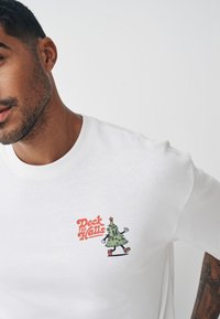 Balta kokvilnas T-krekls ar zaļu multfilmas Ziemassvētku egles attēlu, sarkanu zvaigzni un krāsainām rotājumiem, kā arī tekstu "Deck the Halls".