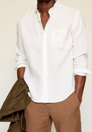 Homme portant une chemise blanche à boutons avec les manches retroussées, un pantalon marron et tenant une veste vert olive dans une main.