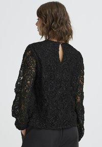 Crna čipkasta bluzica s složenim cvjetnim uzorcima, dugim rukavima i detaljem u obliku ključanice na leđima. Mekana tekstura i uski kroj.