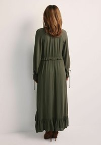 Długa oliwkowa sukienka z falbaną na dole, elastycznym stanem i długimi rękawami z wiązaniami przy nadgarstkach. Gładka tekstura materiału.