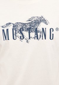 Postarzona granatowa grafika galopującego konia zintegrowana ze słowem "MUSTANG" na jasno beżowym tle tkaniny.