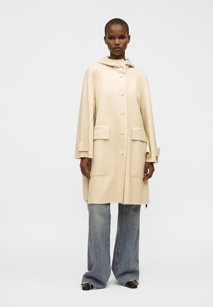 Mujer con un abrigo capa color crema con botones y grandes bolsillos, vaqueros azules de pierna ancha y zapatos negros, de pie frente a un fondo blanco liso.