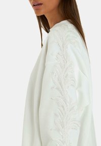 Felpa bianca con ricamo a foglia in paillettes testurizzate lungo le maniche. Tessuto morbido con una vestibilità rilassata e scollatura rotonda.