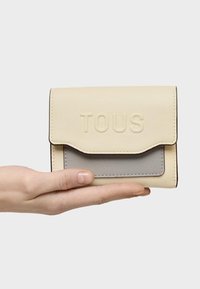 Portefeuille synthétique jaune avec accent gris, finition texturée, logo "TOUS" embossé, et rabat pliable, tenu dans une main sur fond blanc.