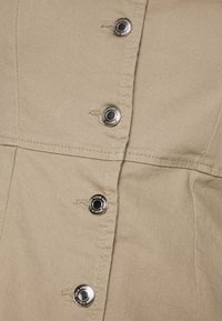 Khaki-kangasmekko, jossa on tiivis vartalo, etupainikkeet metalliosilla, ompeluyksityiskohtia ja sileä kangas.