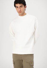 White sweatshirt with a ribbed neckline and hem, featuring long sleeves. Pairing with khaki cargo pants with side pockets and yellow logo accent.